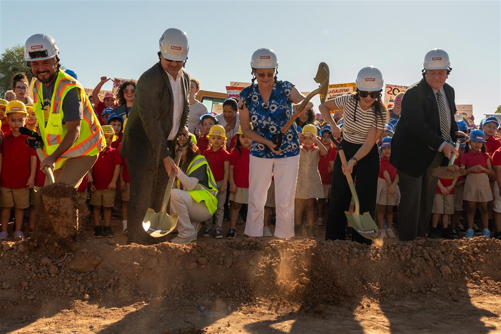 People dressed in hard hats and safety vests use shovels to dig into the ground